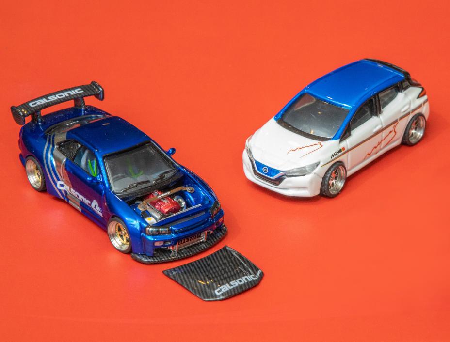 Duas miniaturas de carros sobre fundo vermelho: à esquerda, um esportivo azul com o capô removido exibindo o motor e a palavra "Calsonic"; à direita, um hatch branco e azul com adesivos nas laterais