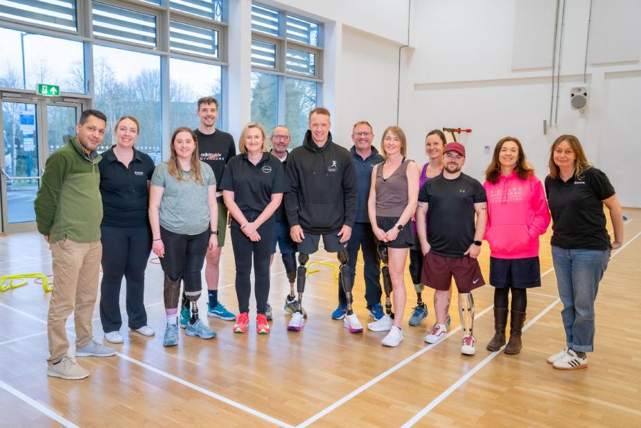 A group photo showing the four new beneficiaries of the Richard Whitehead Foundation Beneficiaries Programme wearing their new sports prosthetics while standing in a sports hall with Richard Whitehead and other team members.