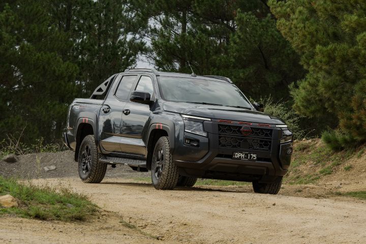 2026 Nissan Navara PRO-4X finished in Boulder Grey shown front three‑quarter on a light gravel track with red grille bars, black wheels, step and roof rails beneath trees.