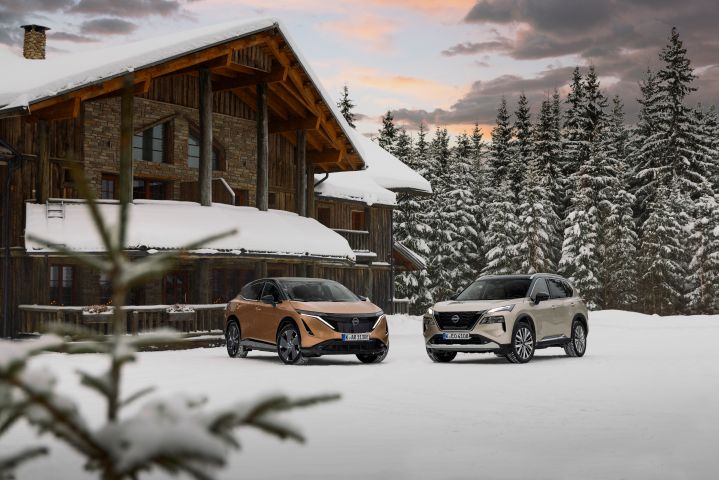 Elektroauto Nissan Ariya e‑4ORCE in Akatsuki Copper mit Dach und Außenspiegel in Pearl Black und silberner Nissan X‑Trail e‑Power e‑4ORCE stehen nebeneinander im Schnee vor Holzhaus und verschneitem Wald im Hintergrund, 3/4 Front