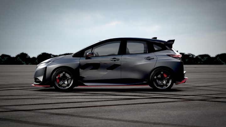A silver and black two-tone sports car, the Aura Nismo RS Concept, parked on an outdoor skid pad in a full side view. The vehicle features a low stance and a large rear wing, with black wheels and visible red brake calipers. The side displays a light gray  'RS,' and a red accent line runs along the lower body. The background shows a cloudy sky and distant trees, with tire marks visible on the asphalt surface."
