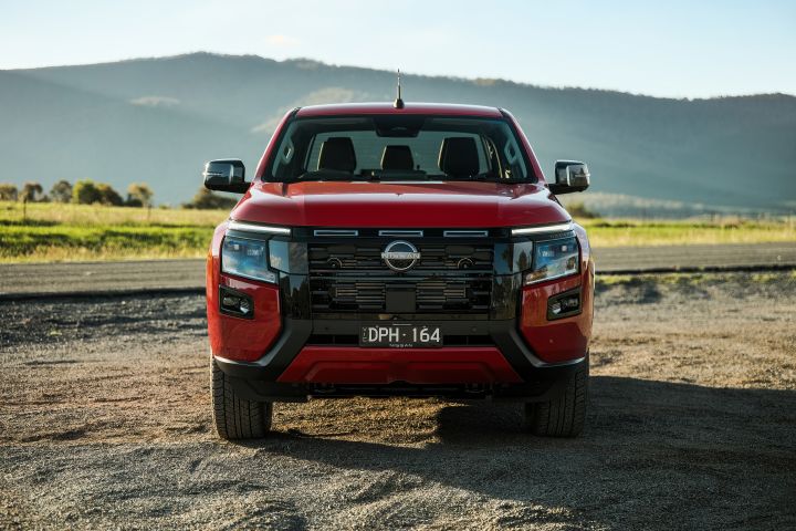 Head-on view of a 2026 Nissan Navara ST-X dual cab in Outback Red, parked on gravel with grassy fields and layered mountain ridges behind the vehicle.