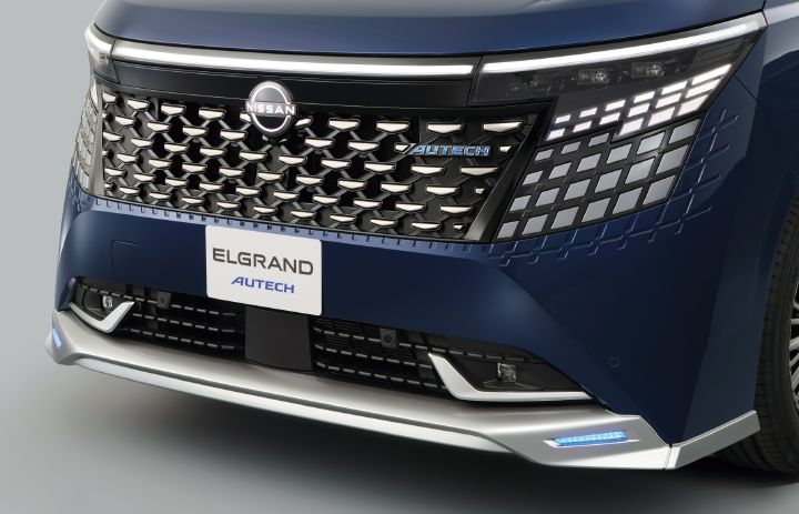 ブルーのエルグランド AUTECHのフロントフェイスを、車両前方やや右側からクローズアップで撮影した画像。大型のドットパターンのフロントグリルが画面中央を占め、中央には日産エンブレム、その右側にはブルーの「AUTECHエンブレム」が配置されている。グリルはシルバーとブラックを組み合わせた立体的なパターンで構成され、高級感と存在感を強調している。
左右のヘッドランプは細長い形状で、デジタル感のある矩形LEDシグネチャーランプが点灯している。フロントバンパー下部にはメタル調フィニッシュのプロテクターが施され、その左右には青白く発光するアクセントイルミネーションが見える。