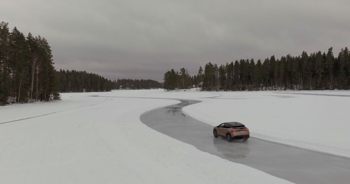 Elektroauto Nissan Ariya e‑4ORCE in Akatsuki Copper mit Dach und Außenspiegel in Pearl Black fährt auf Eissee mit schneebedeckten Bäumen im Hintergrund, 3/4 Heck, Drohnen-Perspektive