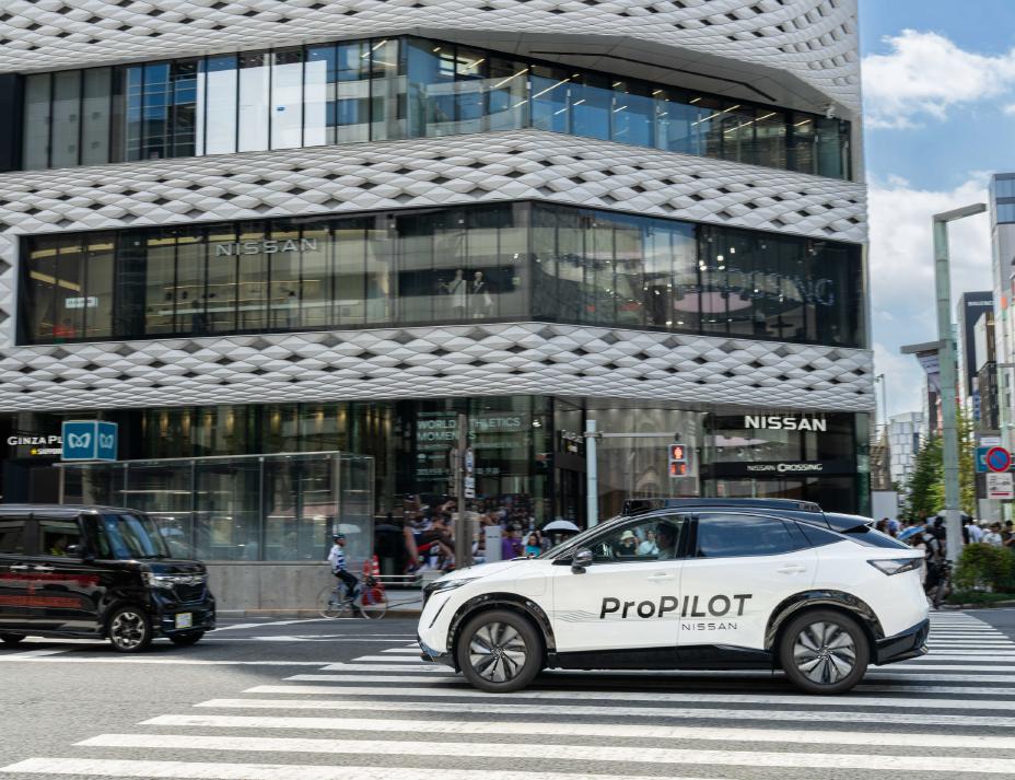 Seitenansicht eines weißen Nissan Ariya, der über einen Fußgängerüberweg fährt und an dessen Seite das Wort „ProPILOT” zu sehen ist. Im Hintergrund steht das moderne Gebäude „NISSAN CROSSING” mit Glasfenstern und einer auffälligen gemusterten Außenwand. Vor dem Gebäude sind Fußgänger und andere Fahrzeuge zu sehen.