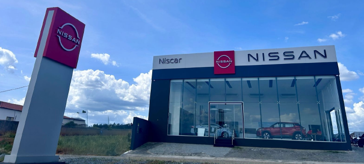 Fachada da nova concessionária Nissan Niscar em Patos, Paraíba. O prédio possui uma estrutura envidraçada com carros expostos no interior. Na frente, há um grande totem vermelho com o logotipo da Nissan. O céu azul com algumas nuvens compõe o fundo da imagem
