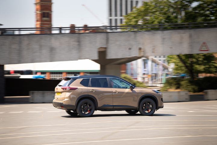 Champagne-coloured Nissan X‑Trail driving through an urban setting, shown in side profile with buildings and an overpass in the background.