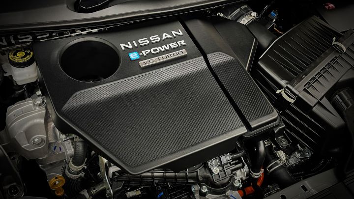 Close-up view of a NISSAN e‑POWER VC TURBO engine. The engine cover is predominantly black with the text 'NISSAN', 'e‑POWER', and 'VC TURBO' visible. Surrounding components include metallic parts and hoses, giving the engine bay a precise and mechanical appearance. The background is dark, highlighting the texture and details of the components.