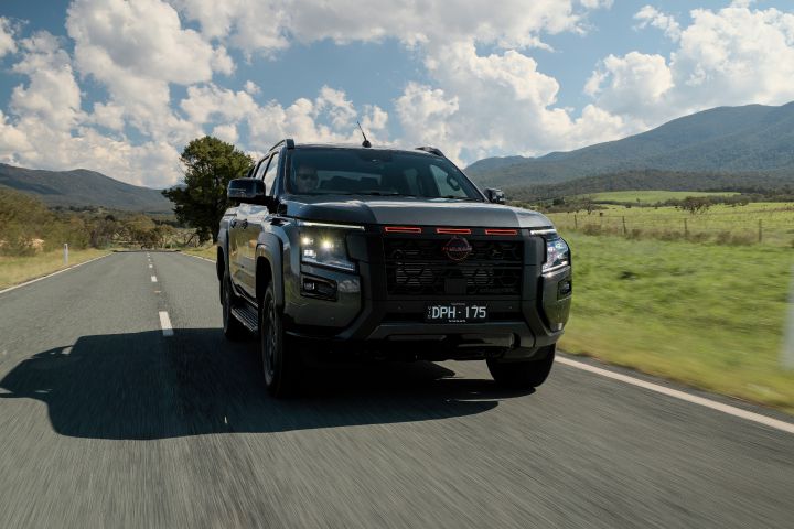 2026 Nissan Navara PRO-4X finished in Boulder Grey presented in a low front-biased driving image with lit daytime running lamps and mountain backdrop.