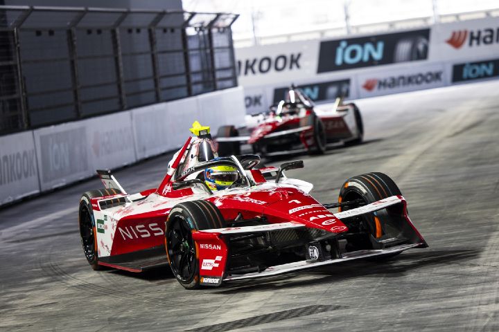 Ein rot-weißer Formel-E-Wagen fährt auf einer Rennstrecke nach links in den Vordergrund. Im Cockpit ist Oliver Roland zu sehen, hinter ihm folgt ein weiterer Formel-E-Wagen. Im Hintergrund sind Sicherheitsbarrieren und Sponsorenschilder zu erkennen.