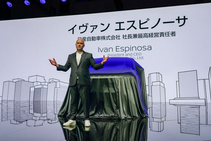 On stage, CEO Ivan Espinosa stands in a suit, gesturing with both hands while delivering a presentation. At the center, a vehicle covered with a black cloth is positioned, and the background screen displays his name and title.