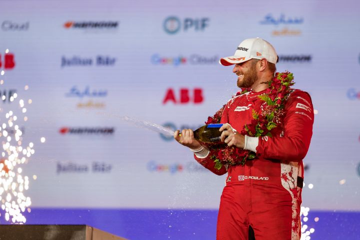 Oliver Rowland vom Nissan Formula E Team spritzt auf dem Podium mit einer Champagnerflasche, während er seinen roten Rennanzug trägt.