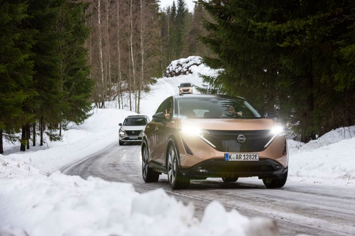 Elektroauto Nissan Ariya e‑4ORCE in Akatsuki Copper mit Dach und Außenspiegel in Pearl Black fährt auf nasser Fahrbahn in verschneiter Landschaft vor Nissan X‑Trail