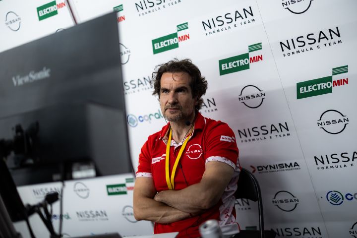 Tommaso Volpe, der ein rotes Nissan Shirt trägt, sitzt mit konzentriertem, nach vorne gerichtetem Blick vor einem weißen Hintergrund, der mit Logos bedeckt ist. Er hat die Arme verschränkt und schaut auf einen Monitor