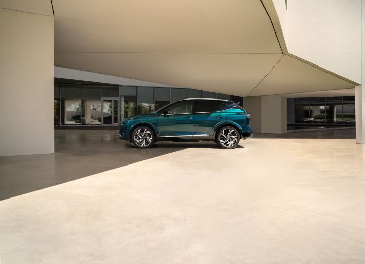 Statische Seitenansicht eines dunkelgrün-blauen Nissan Qashqai mit e‑Power-Antrieb der nächsten Generation vor einem hellen Gebäude mit großzügiger Verglasung