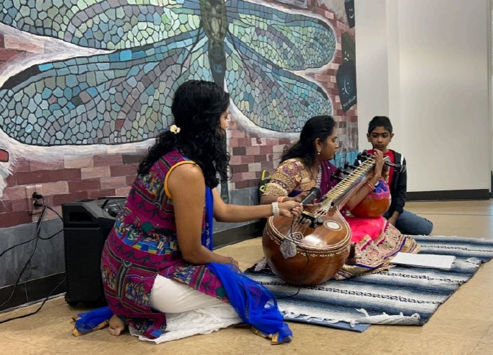 Three women site on the floor playing traditional music, a mosaic wall is behind them.