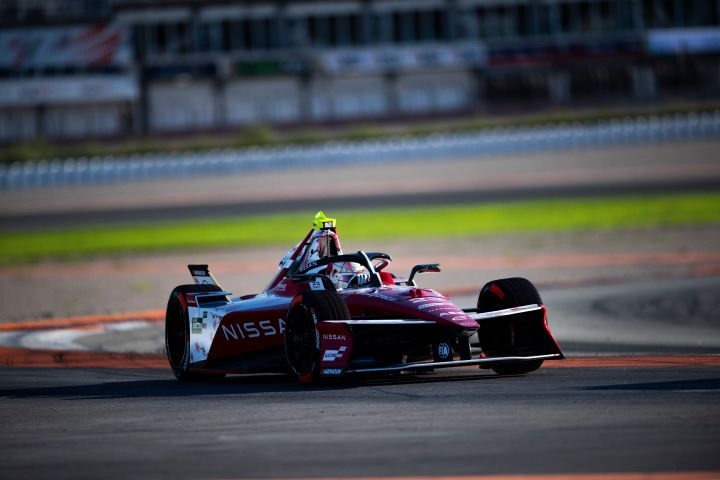 Front on shot of Norman Nato in the #23 car turning left into a corner with a strip of green grass and buildings visible in the background.