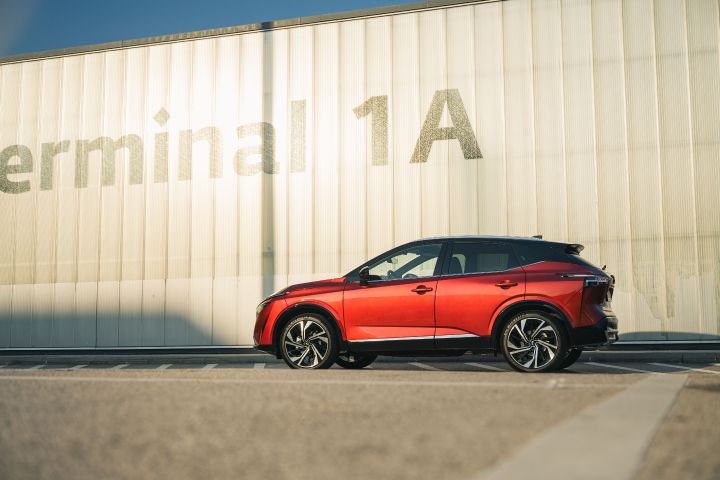 Ein Nissan Qashqai in Fuji Sunset Red steht seitlich vor einem einstöckigen Bauwerk mit der Aufschrift Terminal 1A