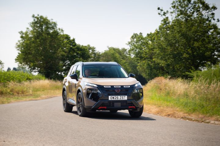 Champagne-coloured Nissan X‑Trail driving along a country road, viewed from the front three-quarter angle, with trees and grassland in the background.