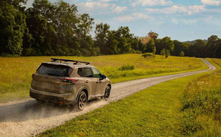 Right rear 3/4 angle of a 2025 Nissan Rogue Rock creek driving on a small dirt road through a field.
