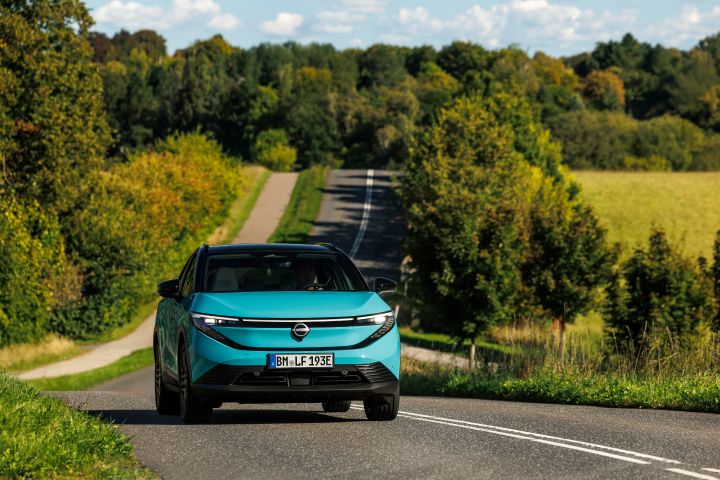 Frontale Fahraufnahme eines türkisen Nissan LEAF auf einer dänischen Landstraße, gesäumt von grünen Bäumen und Wiesen