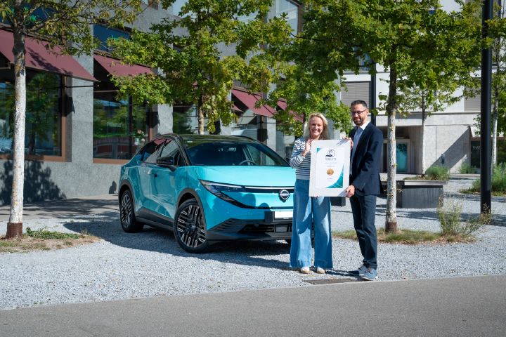 Eine Frau übergibt einem Mann eine große WWCOTY Siegerurkunde, beide stehen lächelnd vor einem neuen Nissan Leaf.