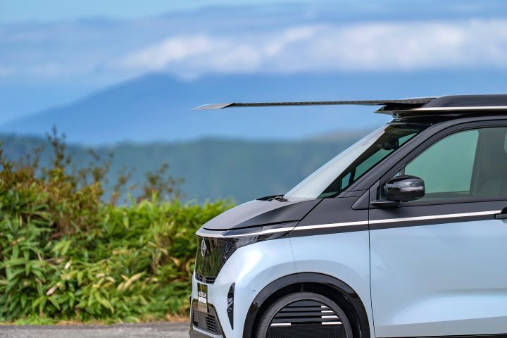 Close-up of the front section of a light blue Nissan SAKURA, facing right, with its solar roof extending prominently forward. The background shows green shrubs, distant blue mountains, and scattered clouds in the sky.