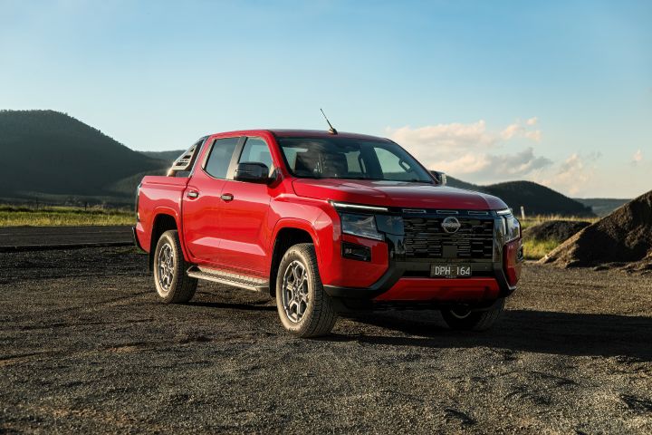 Front three-quarter hero shot of a 2026 Nissan Navara ST-X dual cab in Outback Red, parked on gravel with the sports bar, black grille, alloy wheels, and mountain landscape clearly visible.