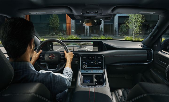 A man in the driver’s seat of a Nissan vehicle, parked in front of a modern house, as viewed from the back seat.