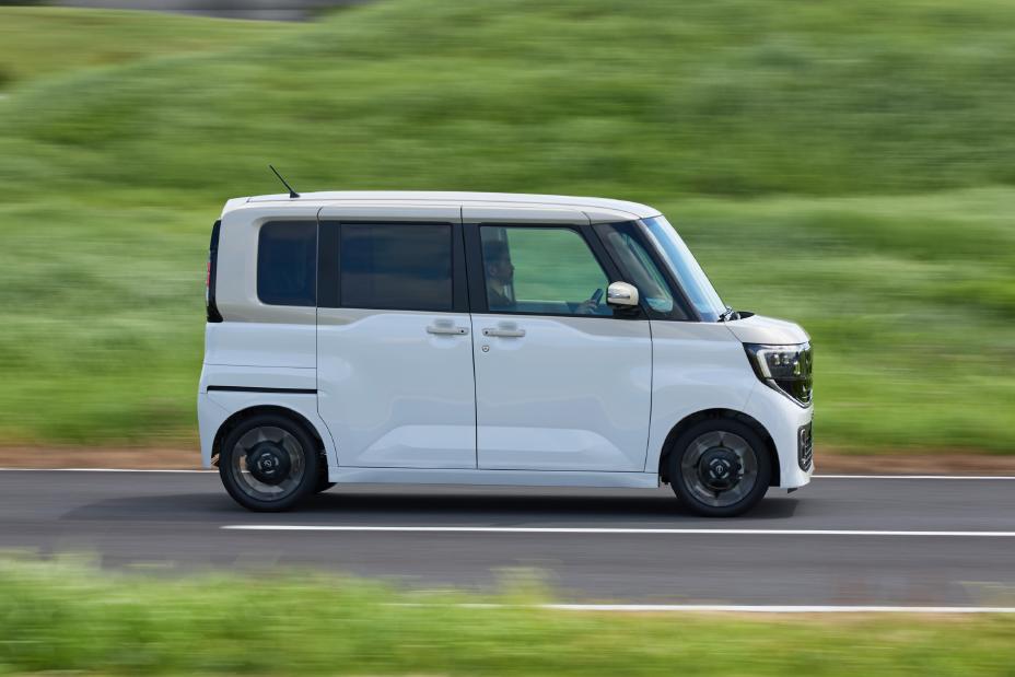 The new Nissan Roox kei car, in white with black wheels and tinted windows, is driving forward on a road with a green background, suggesting motion. The vehicle features a boxy design and is captured in a dynamic scene.