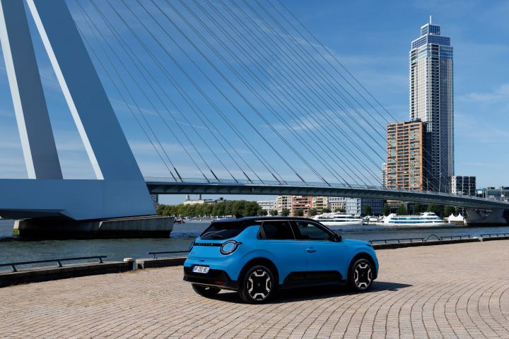 3/4-Heckansicht eines blauen Nissan Micra an einer Kaikante vor einer großen Brücke und einem Hochhaus im Zentrum von Rotterdam