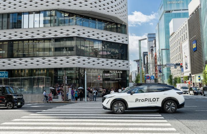 Ein weißer Nissan Ariya fährt über einen Fußgängerüberweg. Im Hintergrund steht das NISSAN CROSSING-Gebäude, das sich durch ein modernes Design mit Glas und weißen Paneelen auszeichnet. Umgeben ist es von Hochhäusern unter einem strahlend blauen Himmel.