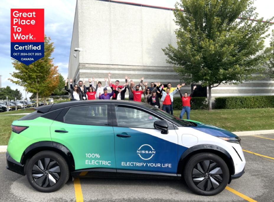 A large group of Nissan employees posing for a group photo in front of a wrapped 2024 Nissan ARIYA and a corporate building in the background