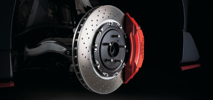 Close-up detail of the high-performance braking system on a Fairlady Z NISMO. With the wheel removed, the image prominently showcases a large ventilated and drilled brake rotor paired with a red multi-piston brake caliper. The caliper features the “NISSAN” logo, while the rotor displays numerous drilled holes designed to enhance cooling performance. Suspension components and the inner wheel well are visible in the background, adding technical detail to the composition. The contrast between the black bodywork and the bright red caliper highlights the performance-focused engineering and motorsport-inspired hardware of the Fairlady Z NISMO.