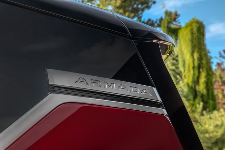 The silver Armada logo at the top of the rear side window on a red 2025 Nissan Armada, trees and blue skies are behind.
