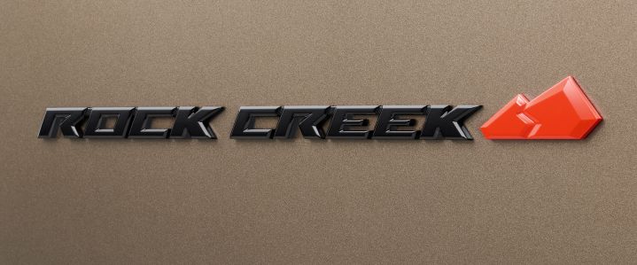 クリスタルブラウンのボディ表面に装着された「ROCK CREEK」専用エンブレムのクローズアップ画像。中央には立体的なブラックの「ROCK CREEK」ロゴが横方向に配置され、その右側には鮮やかなラバレッドの山形モチーフがあしらわれている。