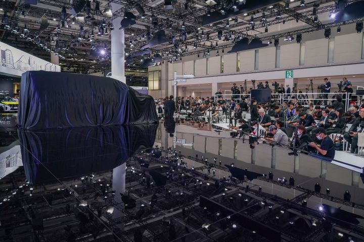 At an exhibition venue, the all -new Elgrand is covered with a black cloth and positioned at the center of the stage.In the background, numerous members of the press are seated, with lighting equipment visible on the ceiling.