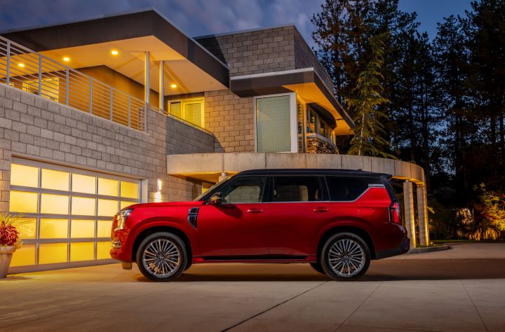 Left profile view of a red 2025 Nissan Armada parked in the driveway of a modern stone mansion at dusk.