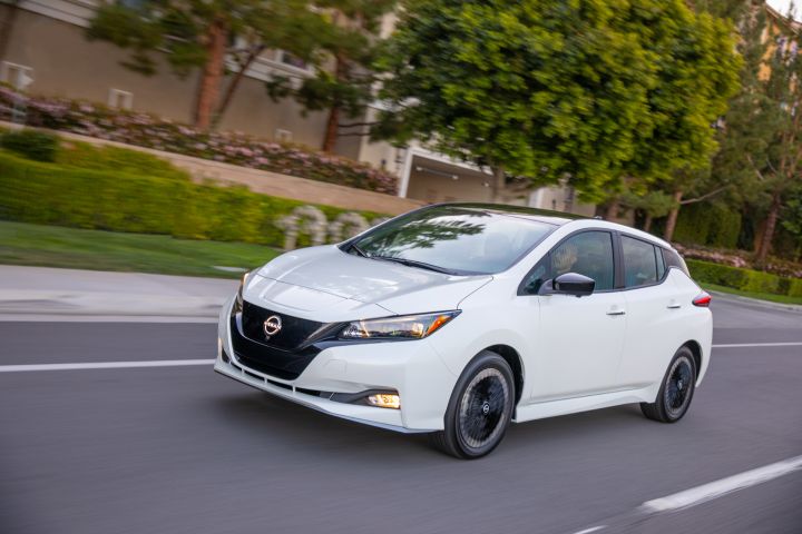 A white 2023 Nissan LEAF driving along a residential road toward the left of the screen.