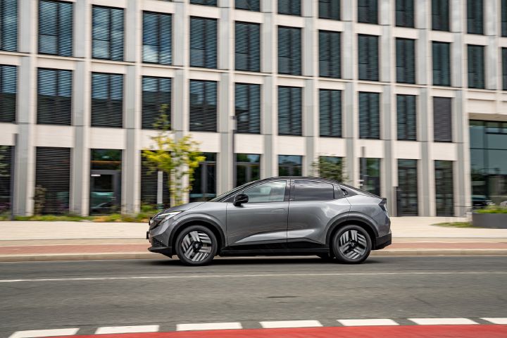 Ein grauer Nissan Leaf fährt seitlich vor einem städtischen Hochhaus über eine Straße