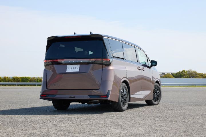 新型エルグランドが、右方向を向いた後方三分の一アングルで静止している。車両は斜め後ろから捉えられ、ブラックルーフと一体化したリアウインドウ、横一文字に伸びるLEDテールランプ、中央の「NISSAN」ロゴが強調されている。周囲には低いガードレールと緑の植生が広がり、その奥には低い建物が見える。上空には淡い青空が広がり、前景には広い灰色の舗装路面が広がる明るい日中の屋外シーンとなっている。