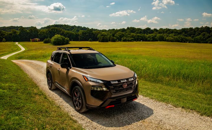 Front right 3/4 angle of a 2025 Nissan Rogue Rock creek parked on a dirt road with field, trees and, blue skies around.