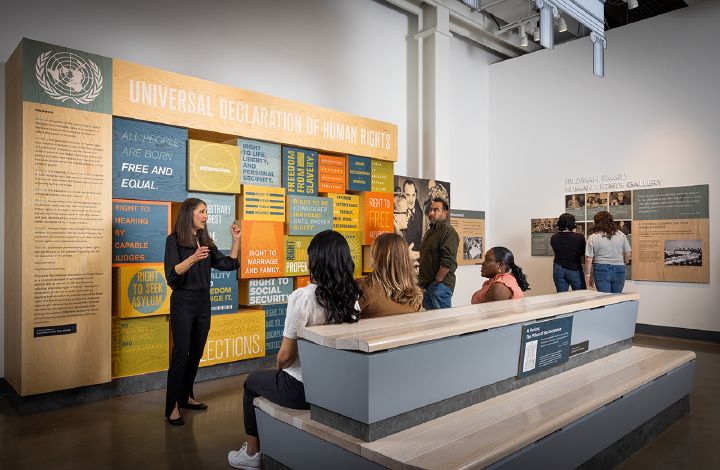 A woman speaking in front of a Human Rights exhibit while a small group of people are sat listening.