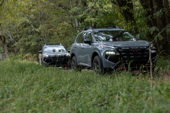 Two Nissan crossovers drive through a grass field in the woods.