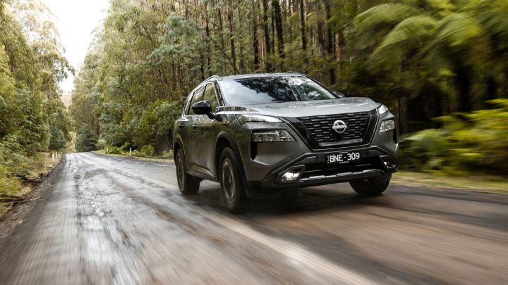 Front ¾ dynamic image of a gray Nissan X‑Trail N-Trek SUV driving through dirt road through a forest