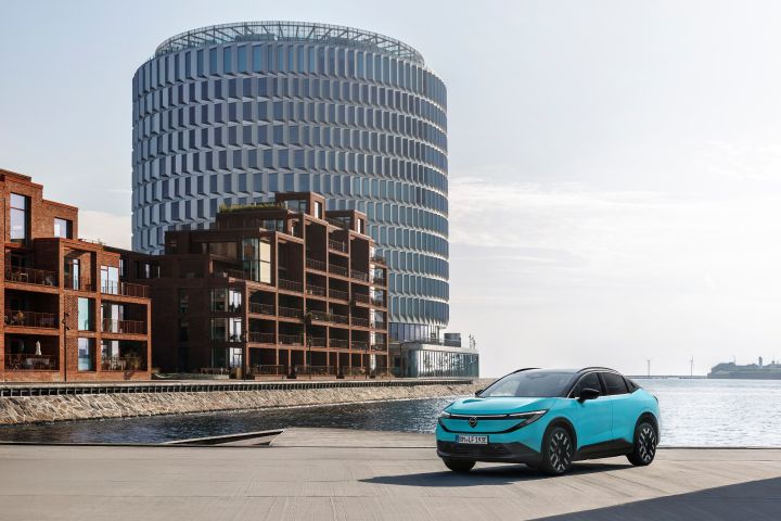 In der rechten unteren Bildecke parkt ein türkisblauer Nissan LEAF, der Rest des Bildes ist gefüllt mit Meer im Hintergrund und modernen Gebäuden auf der linken Seite