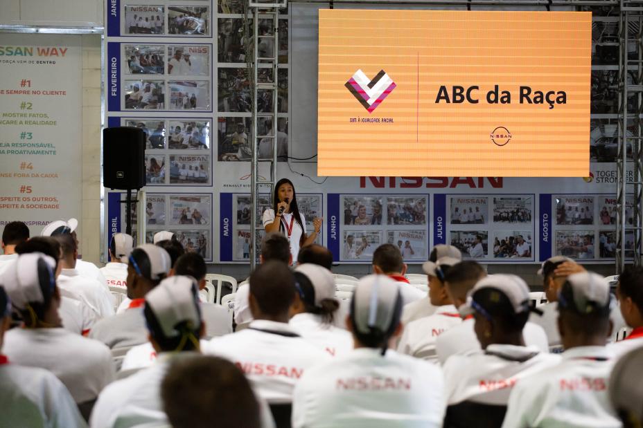 Imagem de funcionária da Nissan no palco na abertura do ABC na Raça na fábrica de Resende com muitos operários da Nissan de costas. Na camiseta deles, a logo da Nissan.