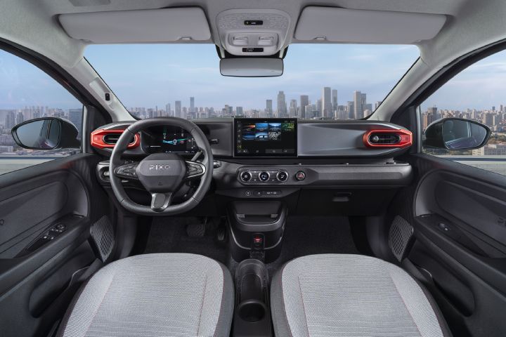 Photograph showcasing Nissan PIXO interior with steering wheel, digital instrument cluster, central touchscreen, climate controls, and textured seats. Red-accented air vents, black dashboard trim, compact center console, and city skyline visible through windshield emphasize vehicle design and urban driving context.