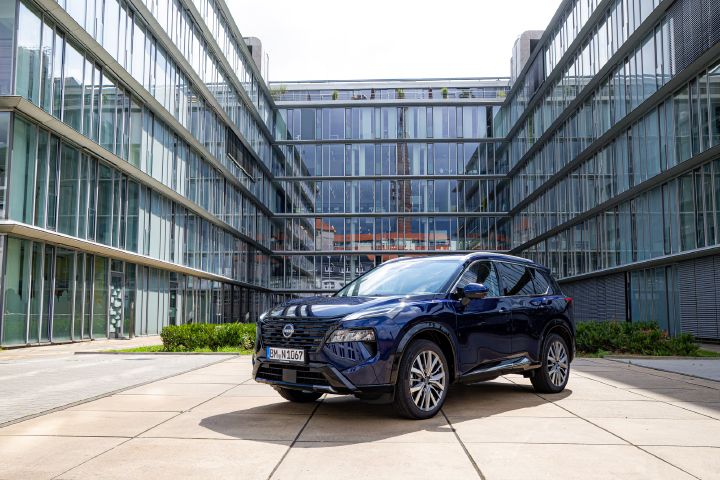 3/4 Frontansicht eines dunkelblauen Nissan X‑Trail, der auf einem Platz steht, der von drei Seiten durch Bürogebäude mit gläserner Fassade umgeben ist