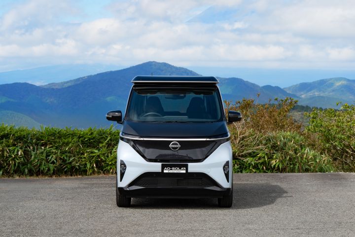 Under a blue sky, a light blue Nissan SAKURA is parked on the road, facing forward. The background includes green shrubs and distant mountains, with white clouds scattered across the sky.